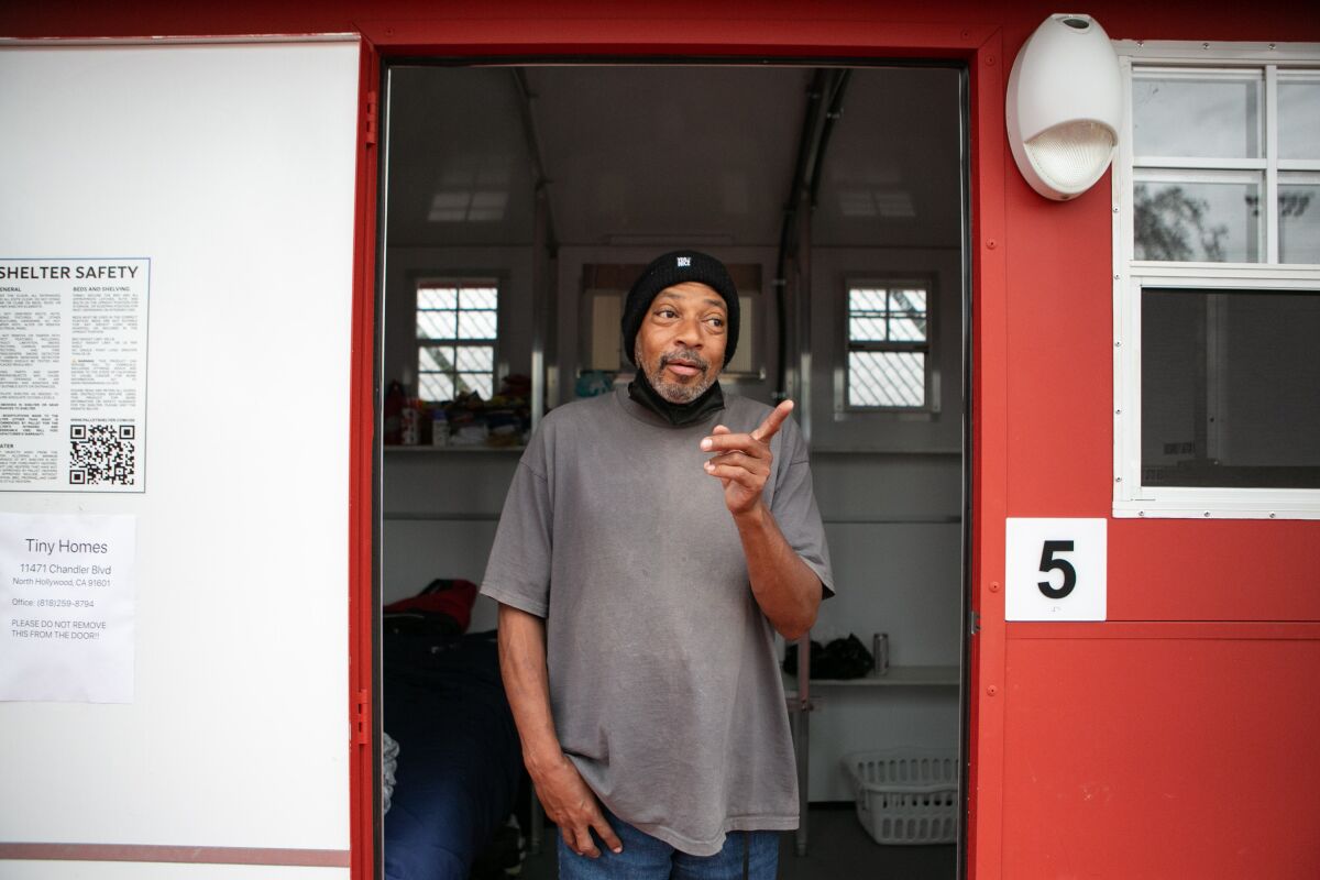 Stephen Smith, in a gray shirt and black ski cap, stands in the doorway of a bright red tiny home