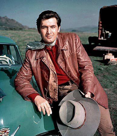 The 6-foot-6 Parker leans on the hood of a vintage car outdoors in a field, wearing a tan leather jacket with a white kerchief around his neck and a cowboy hat in his hand.