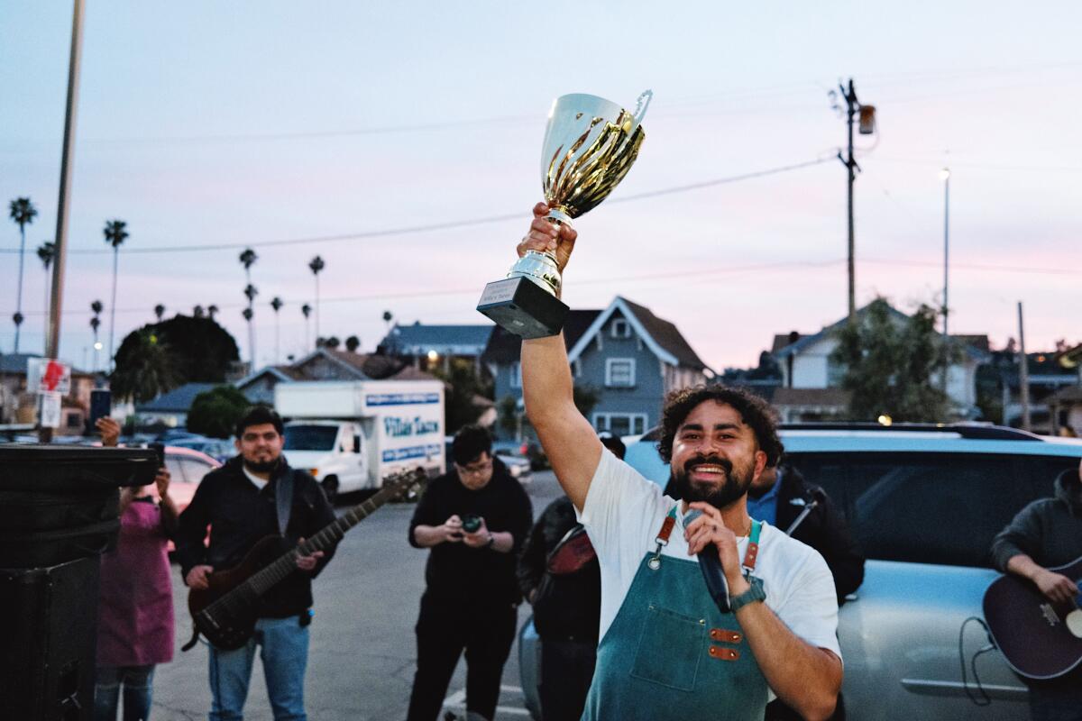 Victor Villa holds a large gold trophy overhead in a parking lot at sundown, with a microphone in the other hand.