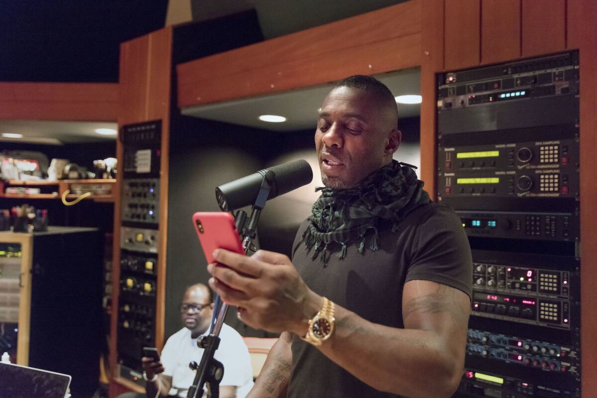 Idris Elba reads lyrics from his phone. Behind him is Poo Bear.
