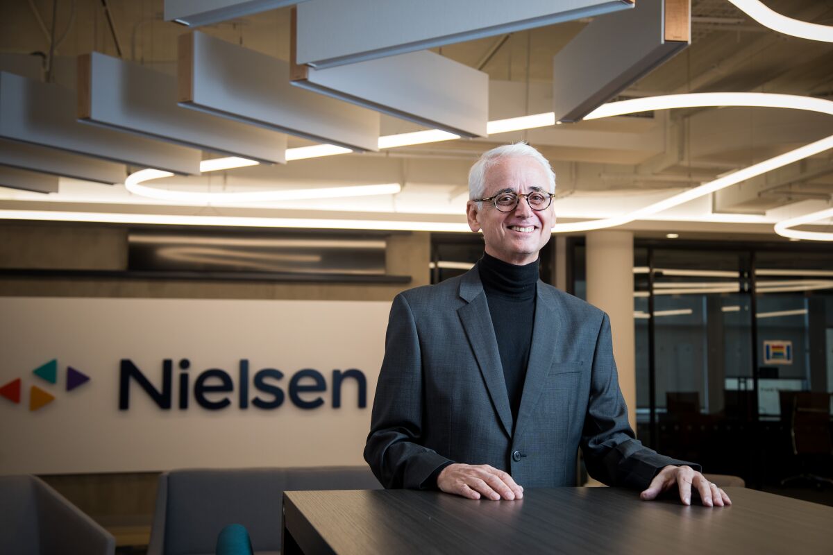 A man in a turtleneck and blazer in front of a Nielsen sign in an office 