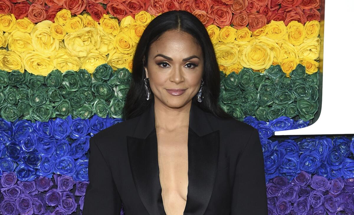 Karen Olivo, in a black suit, stands before a wall of rainbow roses.