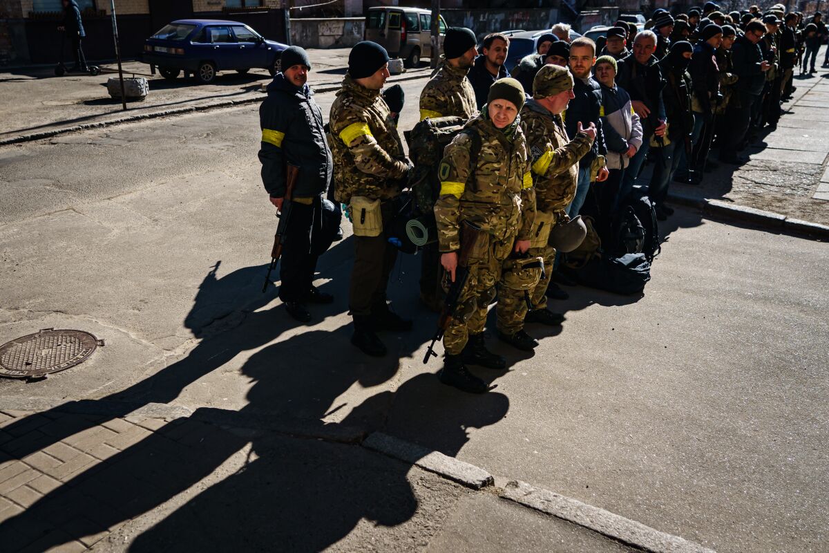 A large group of people in dark and camouflage jackets stand in formation in a street
