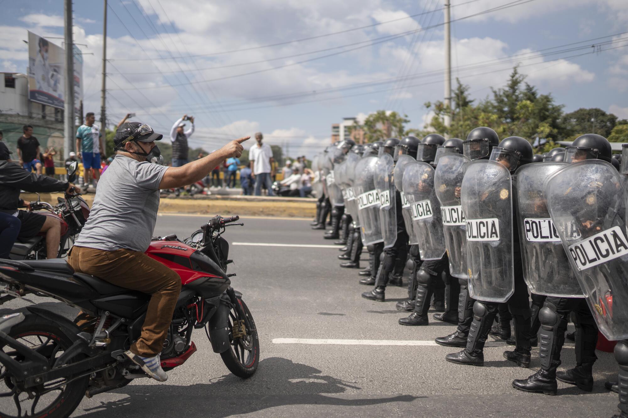 A demonstrator on a motorcycle pointing as he confronts a line of police in riot gear a few feet away as others look on