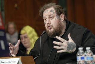 A man with brown hair and multiple face tattoos wearing dark clothing speaks into a microphone in a court room