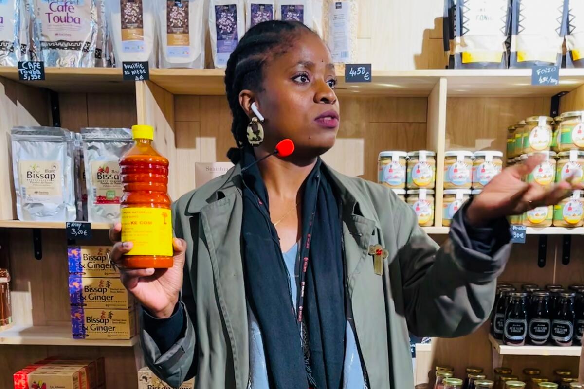 A woman stands in front of shelves with jars and cans and holds up a bottle of liquid.
