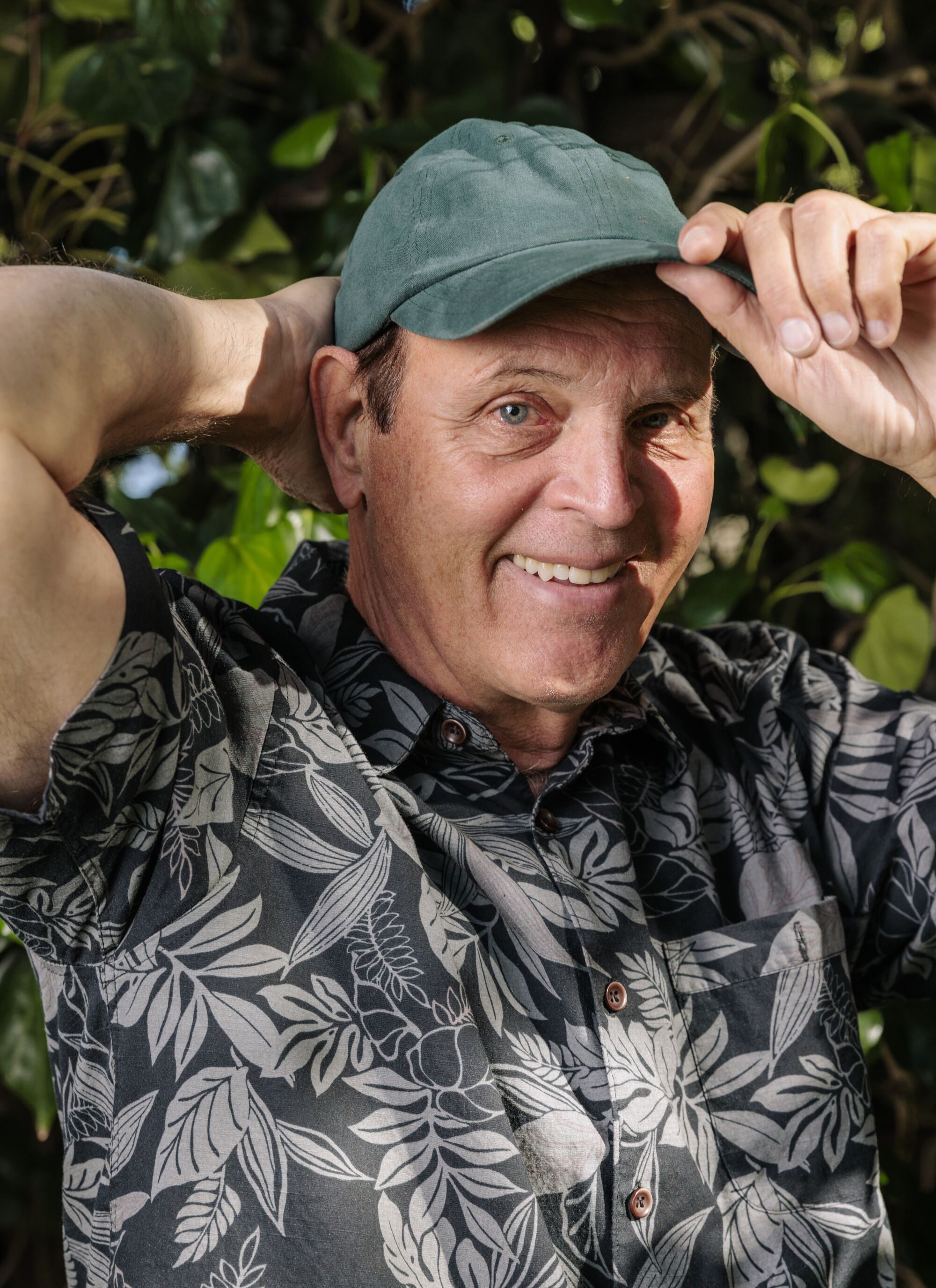 A man in a black floral shirt smiles and tips his cap
