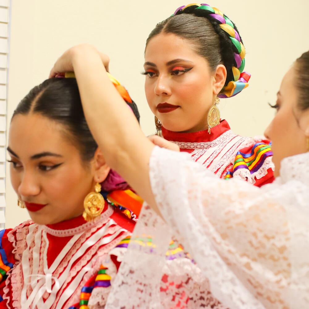 Three young women in folklórico apparel help each other with their hair before a show