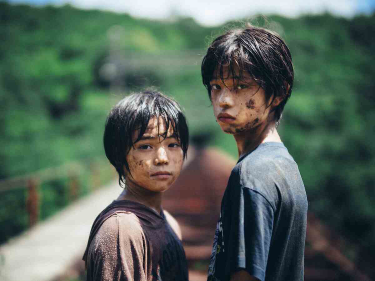 Two young people with dirty, sad-looking faces stand on a road, looking toward the camera 