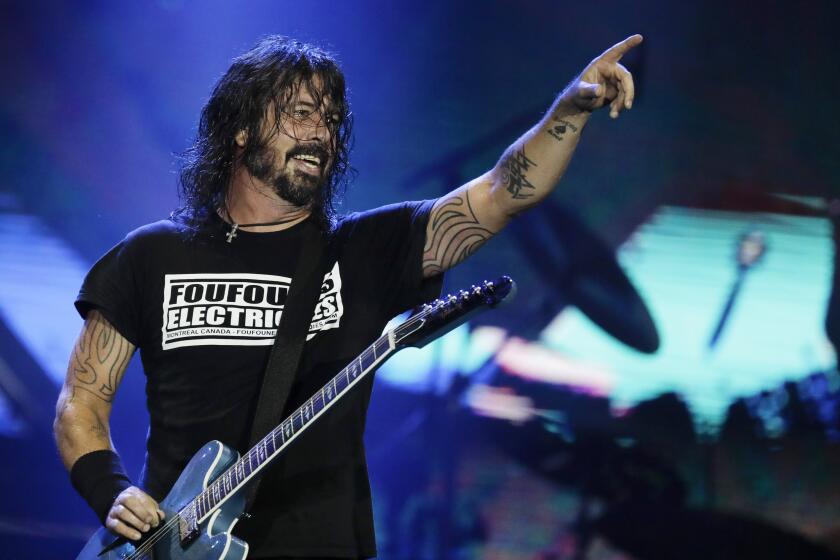 Dave Grohl in a black t-shirt on a stage pointing his left arm in the air and holding a guitar