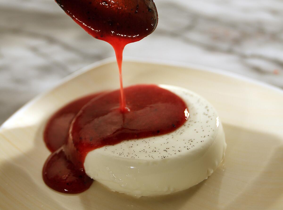 A spoon drizzles a red sauce over a round panna cotta 