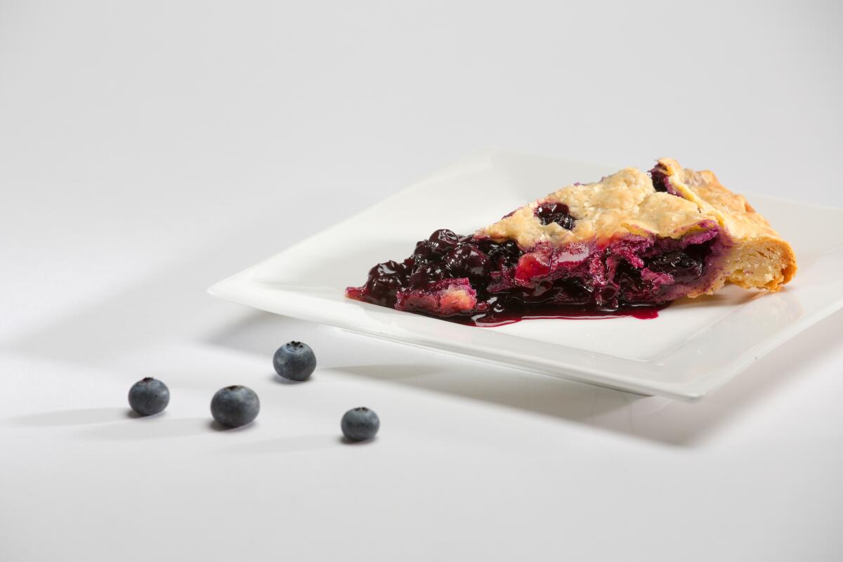 A slice of berry pie on a square white plate with four stray blueberries nearby