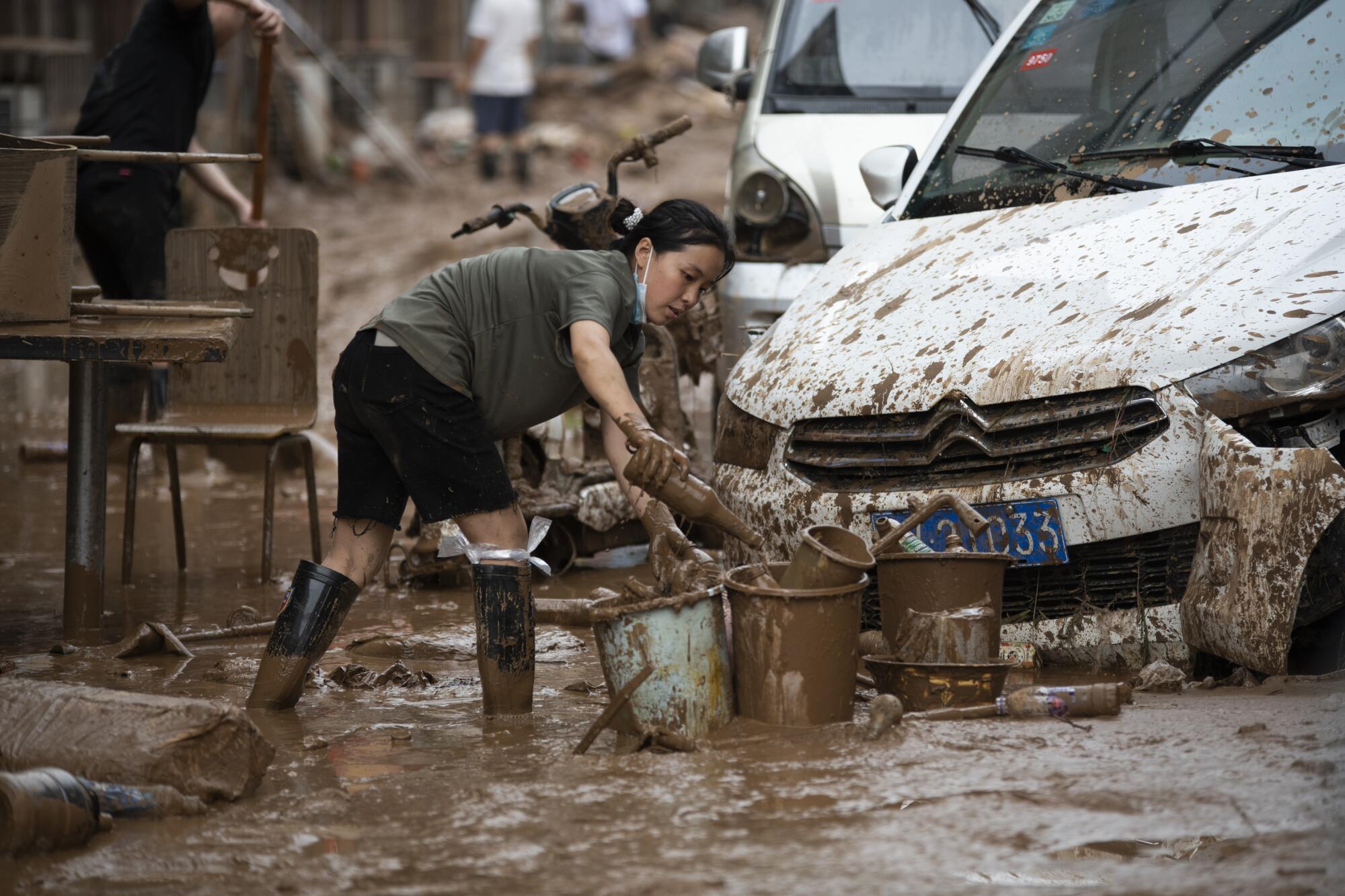 A woman in rain boots holds a muddy bottle as she stands amid mud 