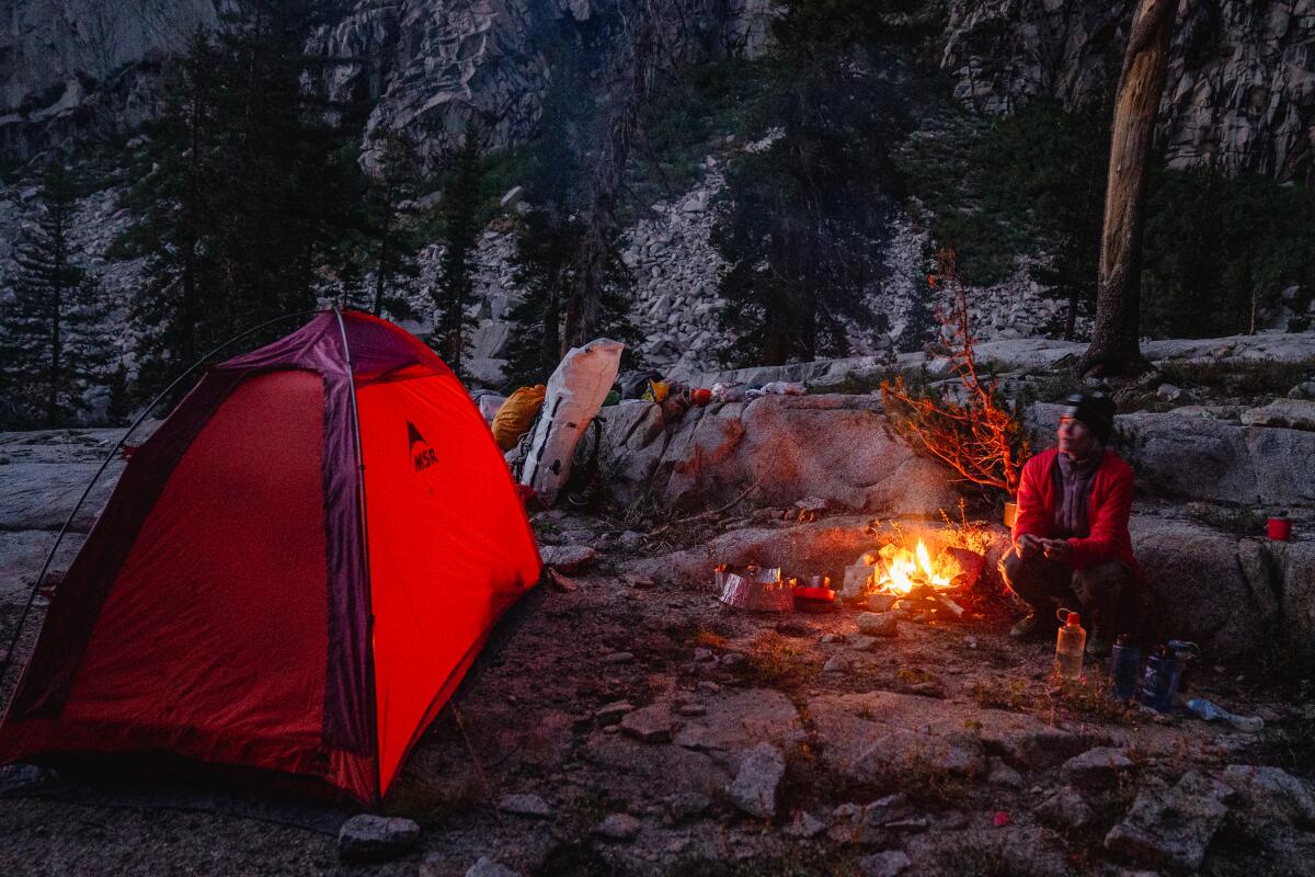 A woman sits near a campfire and tent in the wilderness.