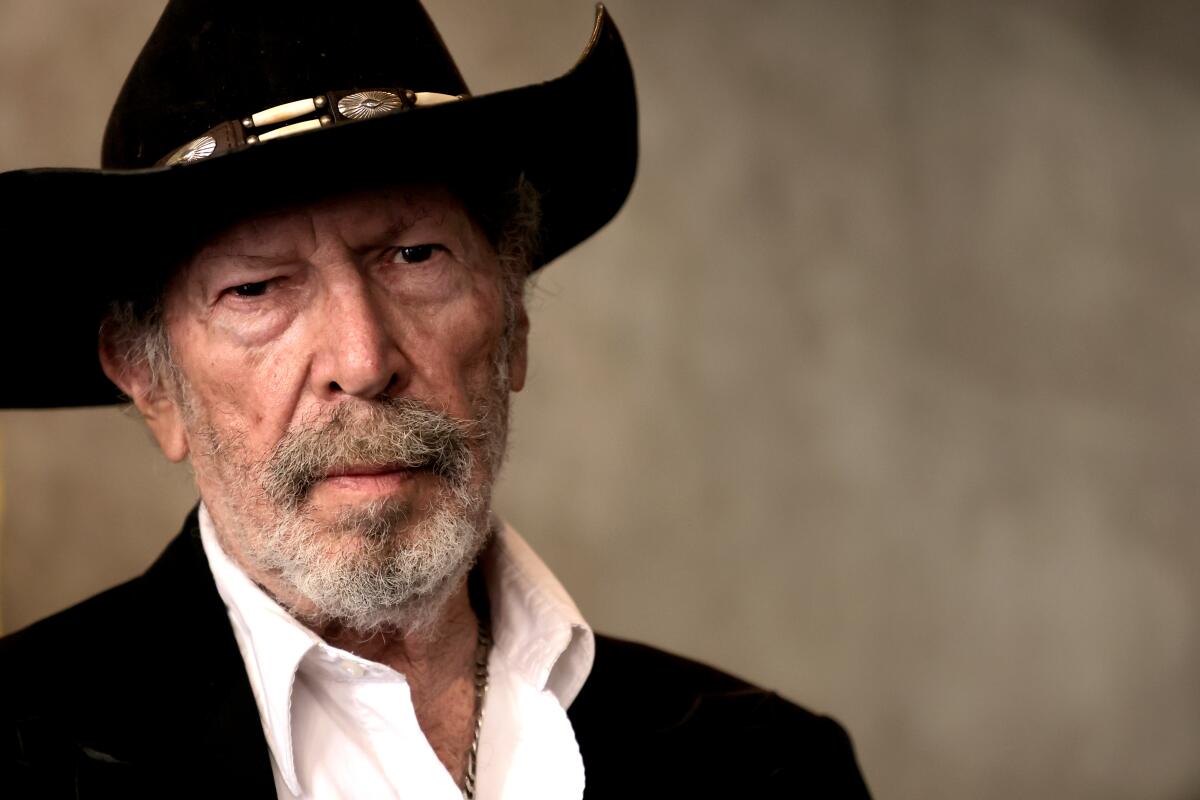 Kinky Friedman wearing a black cowboy hat, black jacket, white open-collar shirt and a serious expression