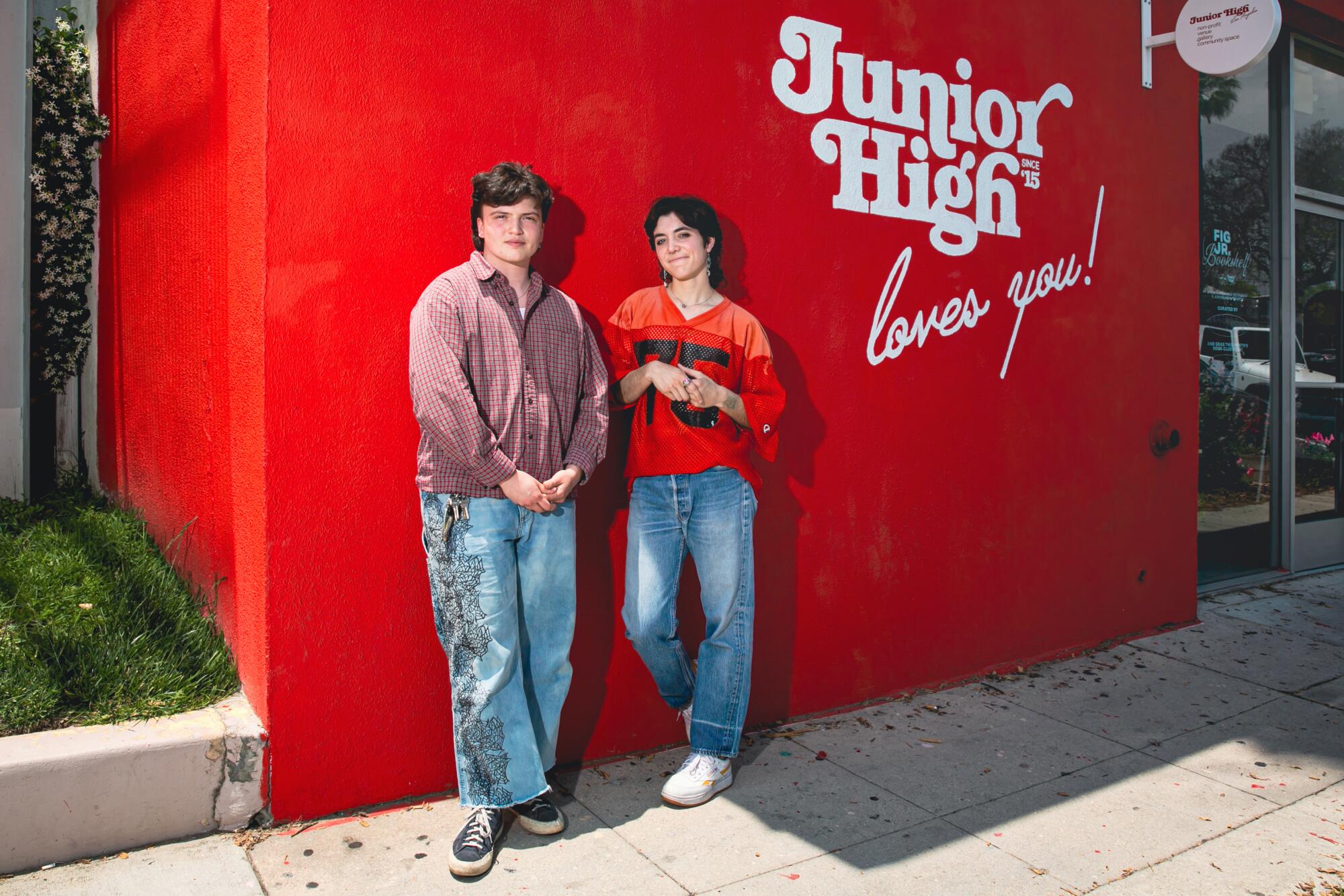 Two people stand for a portrait in front of a red wall with the words "Junior High loves you!"