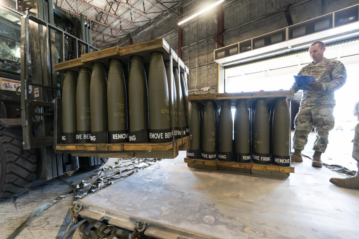 A soldier holding a clipboard oversees loading of pallets of artillery shells onto a shipping platform