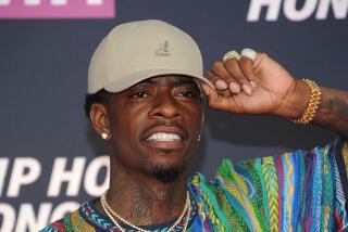 Rich Homie Quan holding the brim of a khaki hat. He smiles and wears a colorful sweater and chains