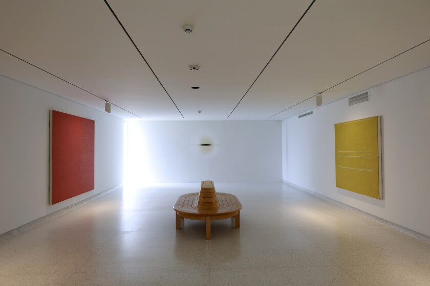 A bench in the center of a museum gallery, with large red and yellow paintings hanging on the walls on either said.