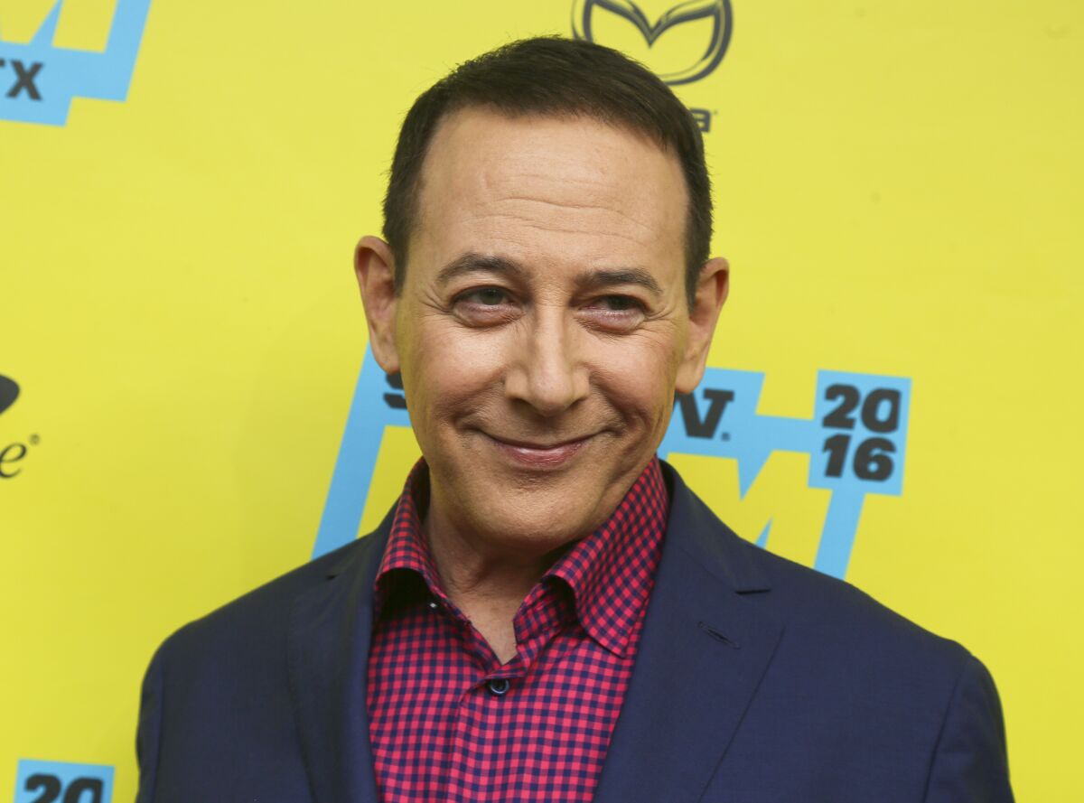 Paul Reubens smiling in a blue suit and checkered red shirt against a yellow backdrop