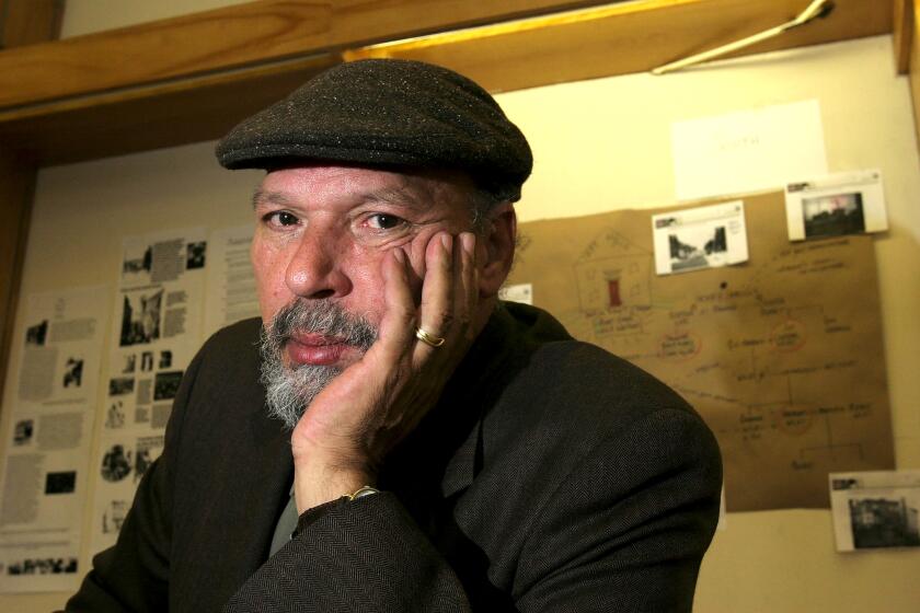 A playwright wearing a new newsboy cap with a salt-and-pepper beard in a black jacket in an office.