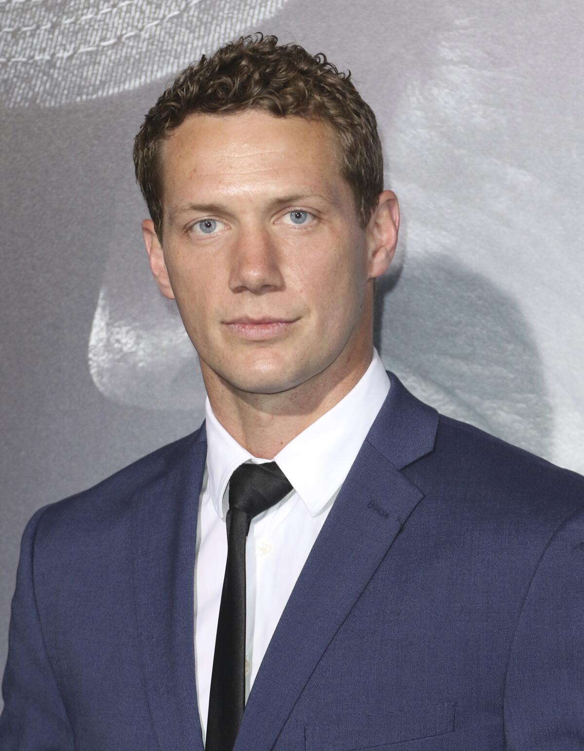 A man in a blue suit, black tie and white dress shirt posing in front of a gray background at a movie premiere