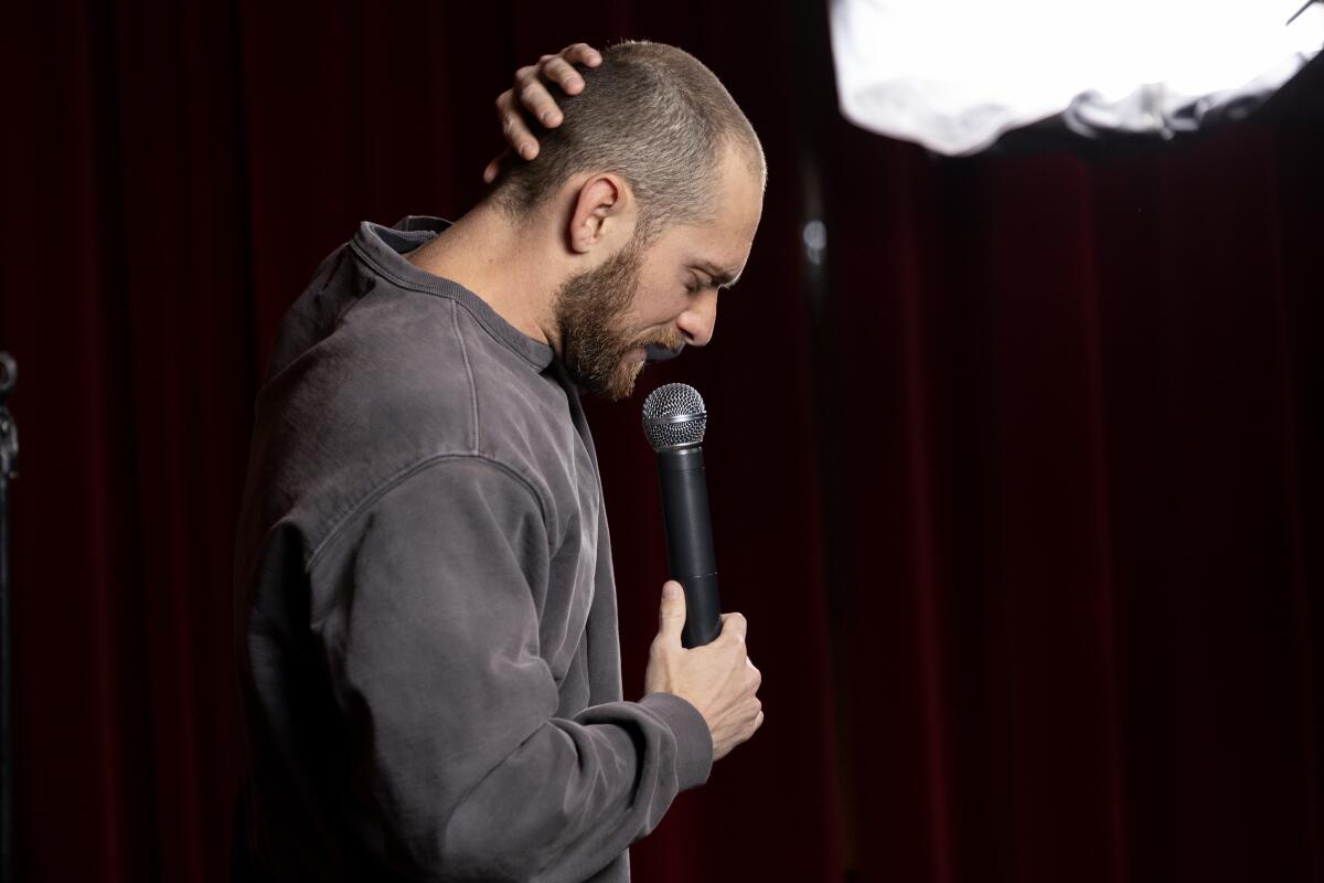 A man rubs his head with one hand and looks down toward a handheld microphone in the other