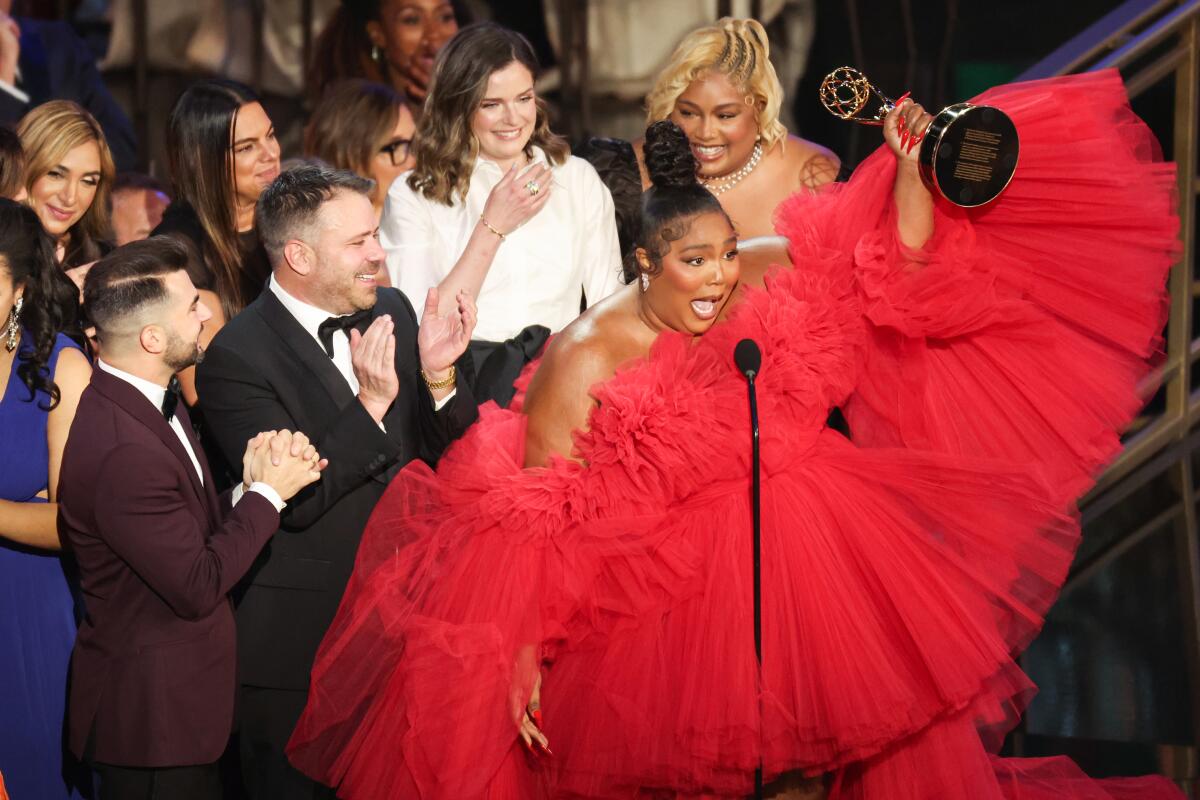 A woman in a red dress speaks into a microphone while holding up a statue. Behind her, people in formal wear clap.