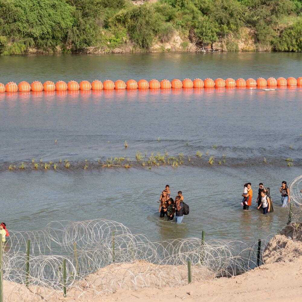 Migrants walk by a string of buoys placed by Texas in the Rio Grande.