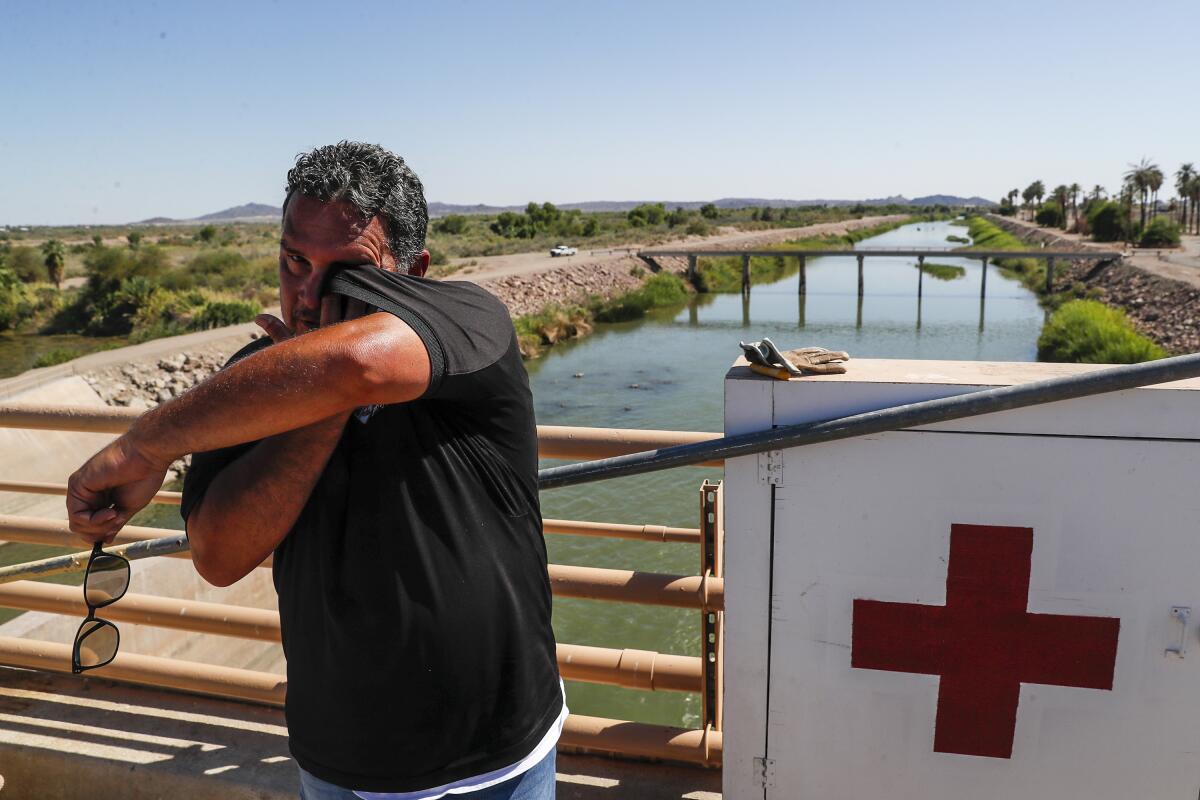 A man uses a shirt sleeve to wipe his eye. He's standing on a bridge over a channel of water.