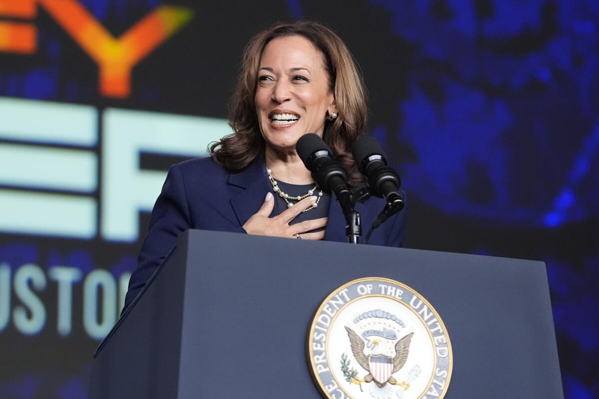 Kamala Harris, smiling with her right hand to her chest, stands at a lectern with large mics and the vice presidential seal.
