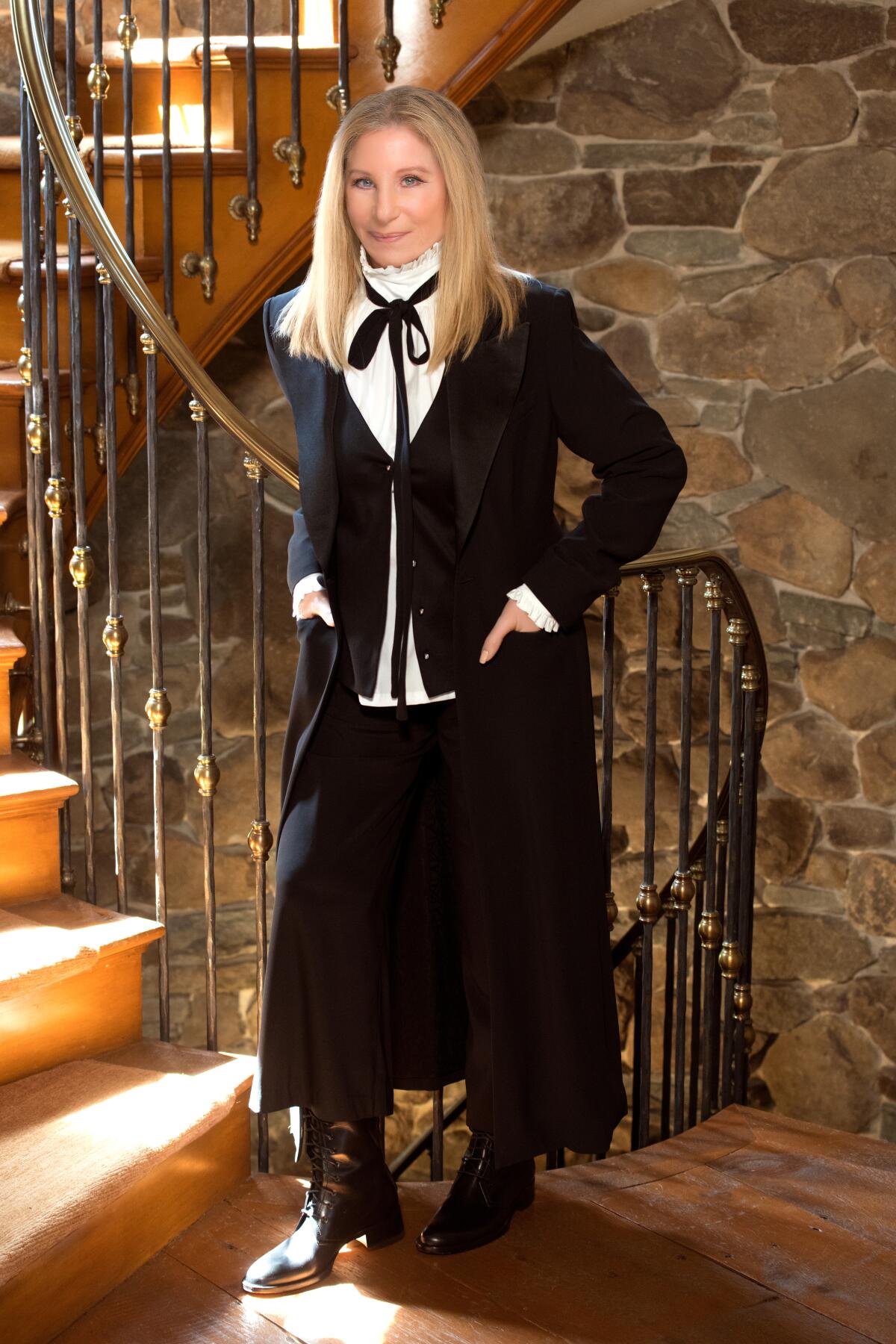 Barbra Streisand wears a black skirt and jacket with a white blouse as she poses at the foot of a staircase.