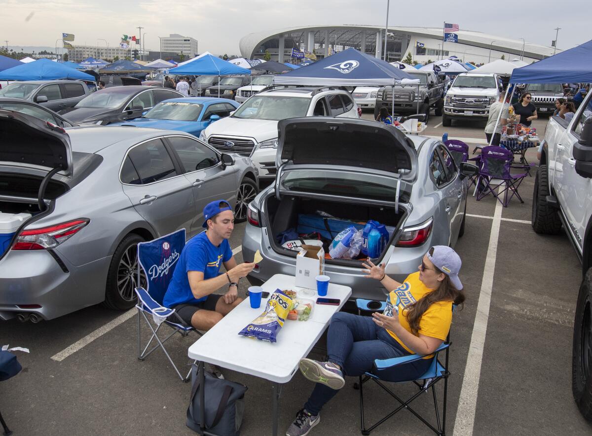 Two people eat and drink at a table behind their car in the parking lot outside SoFi Stadium on Aug. 14, 2021 in Inglewood.