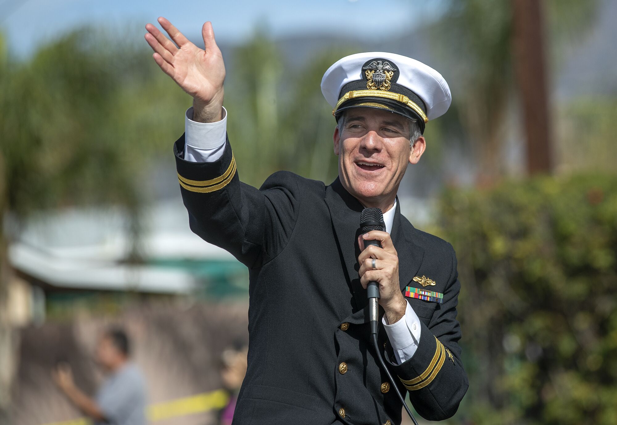 Los Angeles Mayor Eric Garcetti, in uniform, holds a microphone and waves 