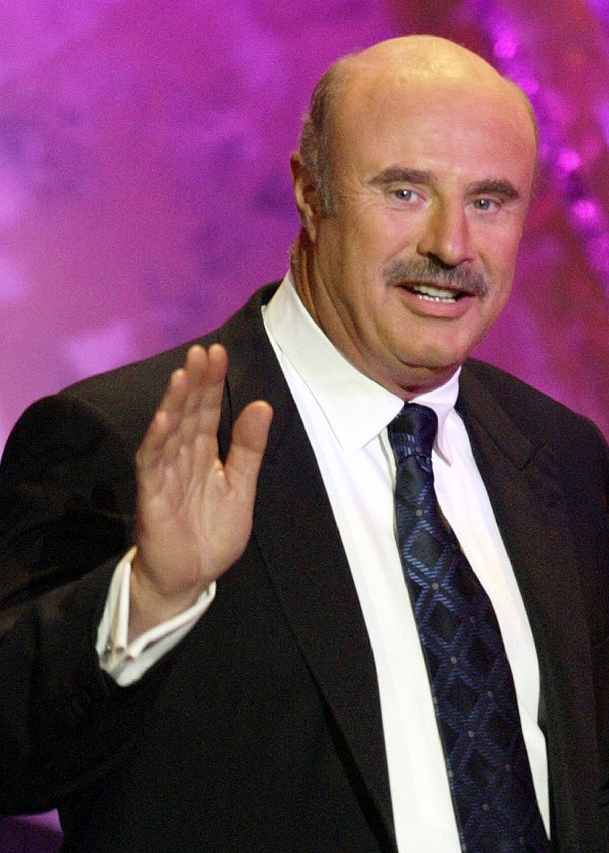 A bald man with a mustache waves with his right hand