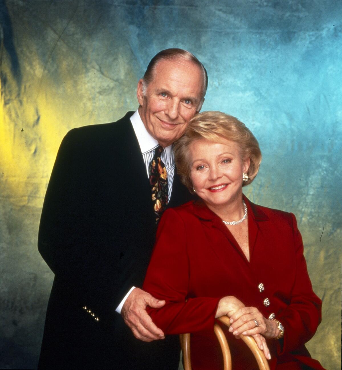A man in a dark suit leans against a woman in a red suit, who is sitting on a chair with her arm resting on top.
