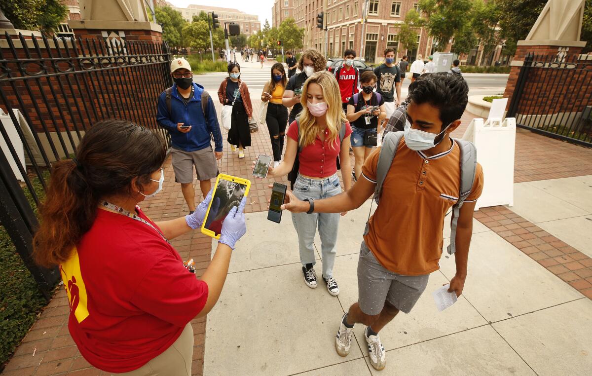 Young people in masks stand in a line at a campus entryway and hold up their phones to a woman.