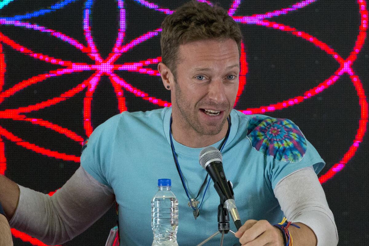 Chris Martin of Coldplay in a light blue T-shirt over a long-sleeved white shirt sitting behind a microphone