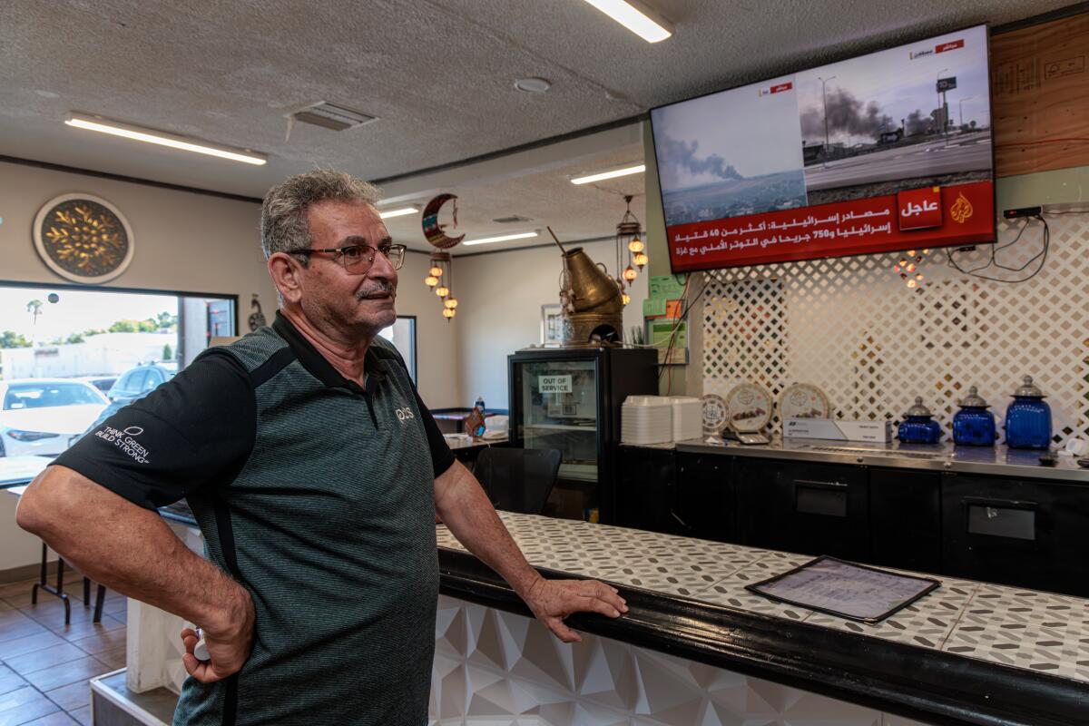 A man stands at a restaurant counter shut to a flat veil TV displaying smoking billowing from land.