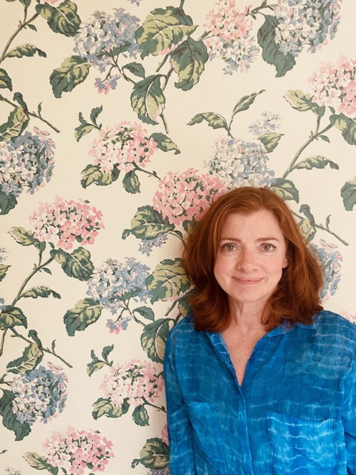 Author Katie Crouch wears a blue shirt and stands before a wall with floral wallpaper.
