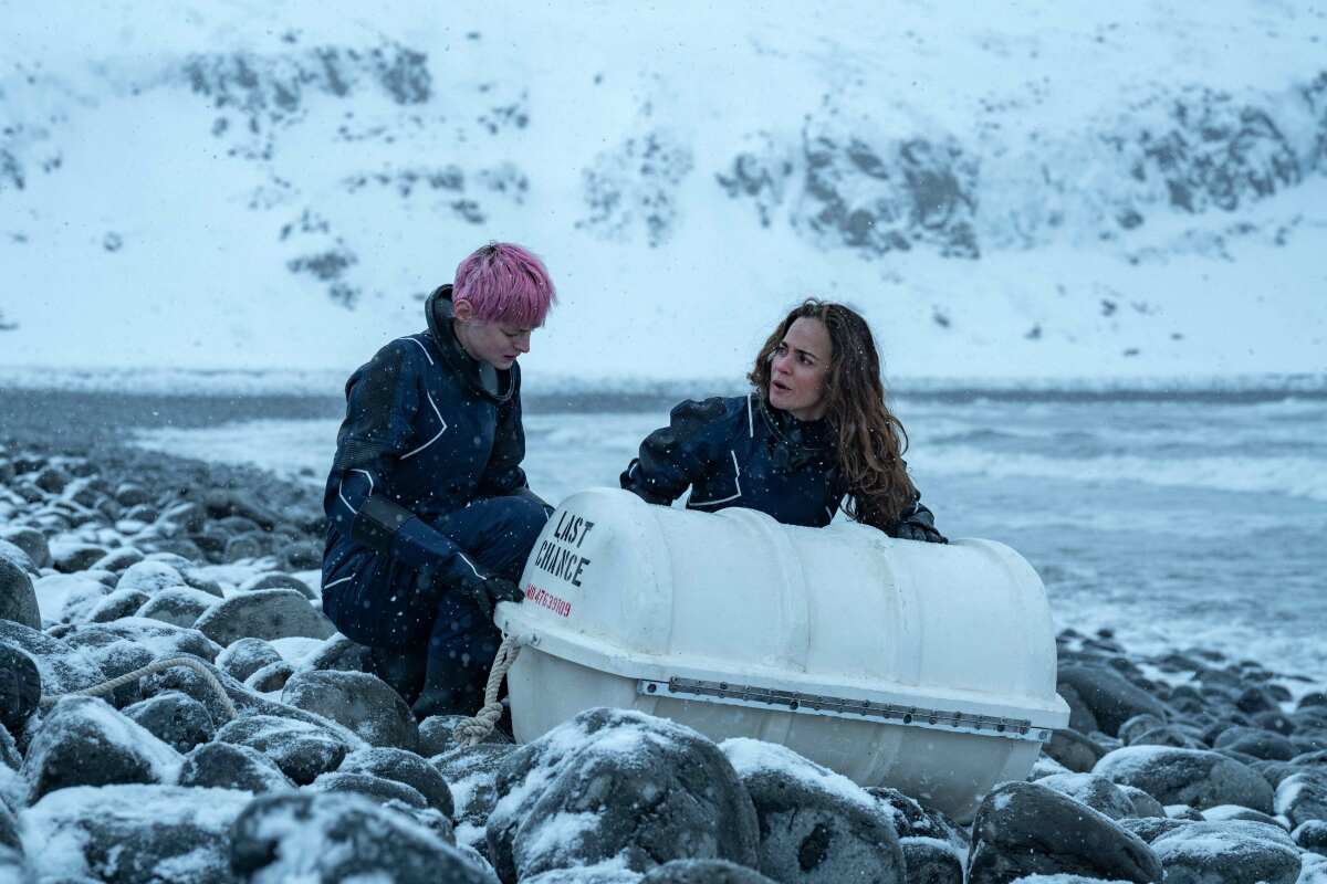 Darby and Sian kneel next to a white capsule labeled Last Chance on a rocky, snowy beach.