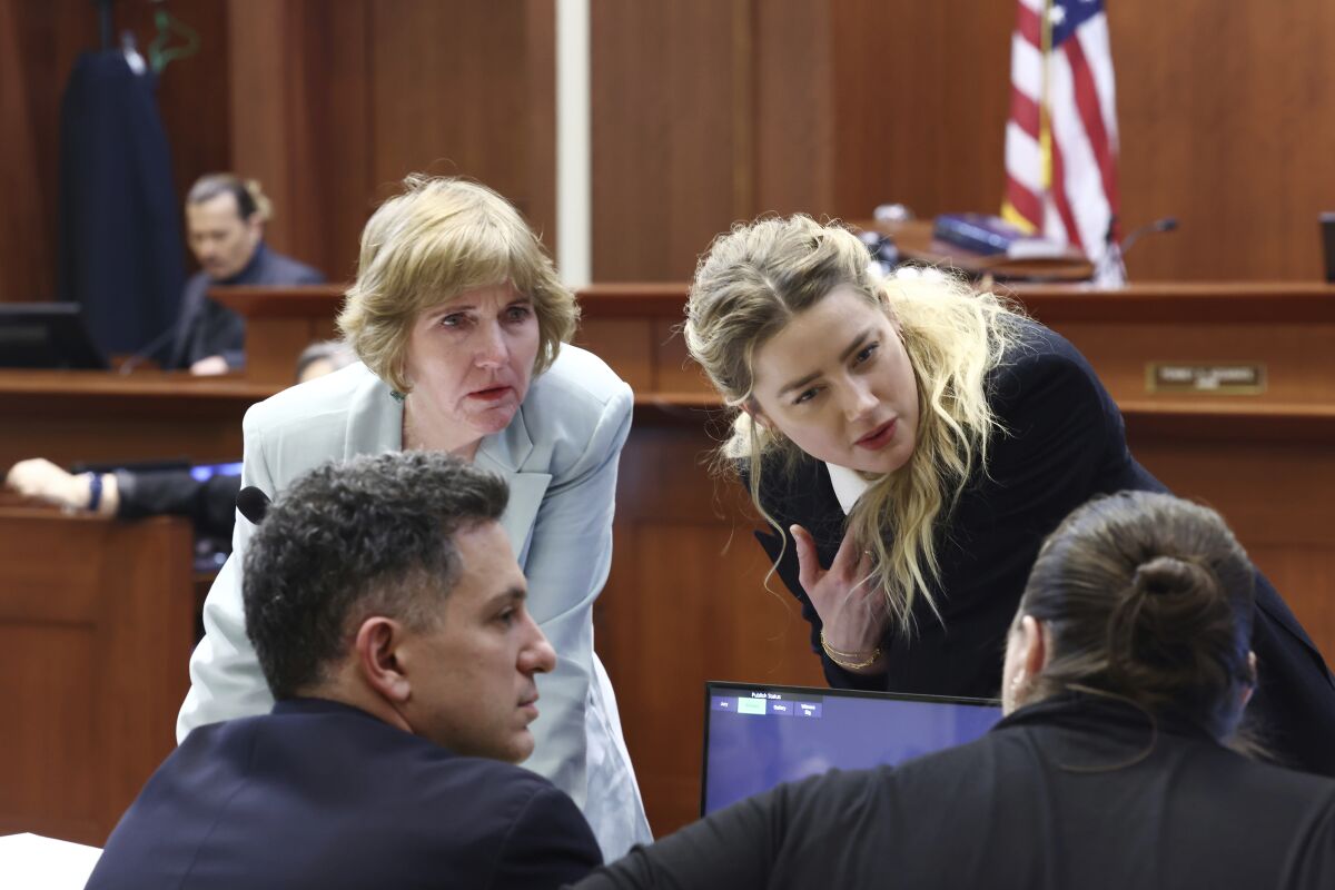A blond woman leans over a desk to speak with three attorneys in a courtroom