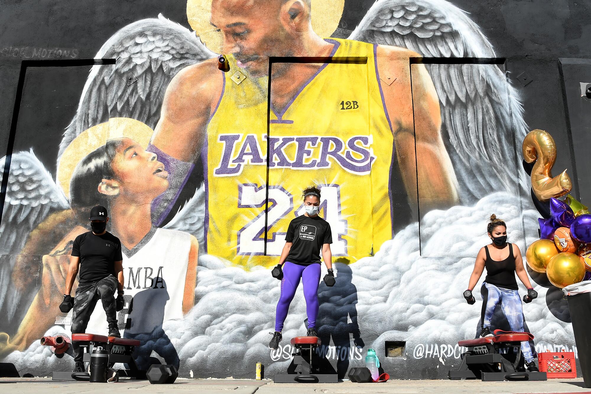 Three people hold dumbbells in front of a mural depicting Kobe and Gianna Bryant with angel wings.