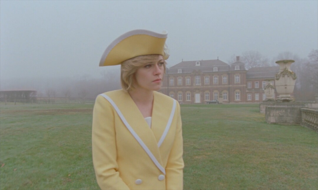 A woman in a yellow dress and tricorn hat stands in the mist outside an English country home.