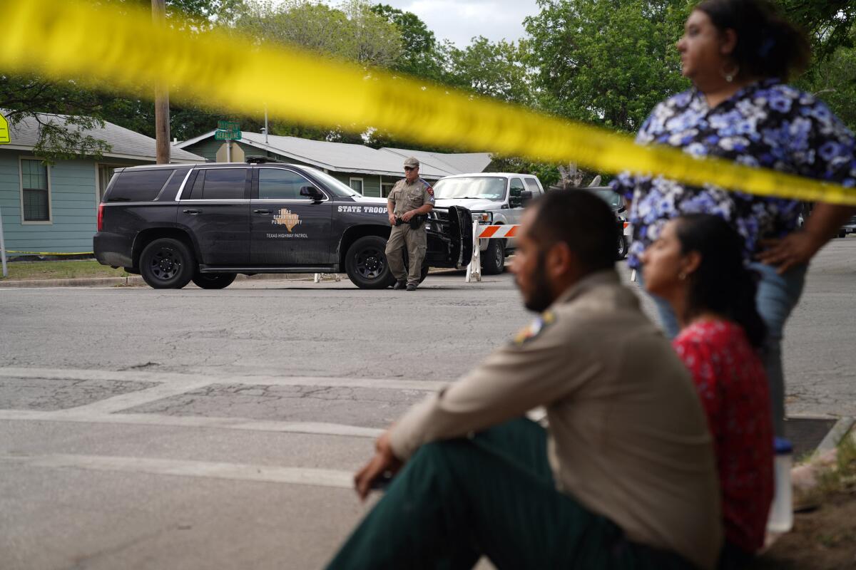 A Texas state trooper stands guard near a patrol SUV while three people wait near yellow barricade tape.