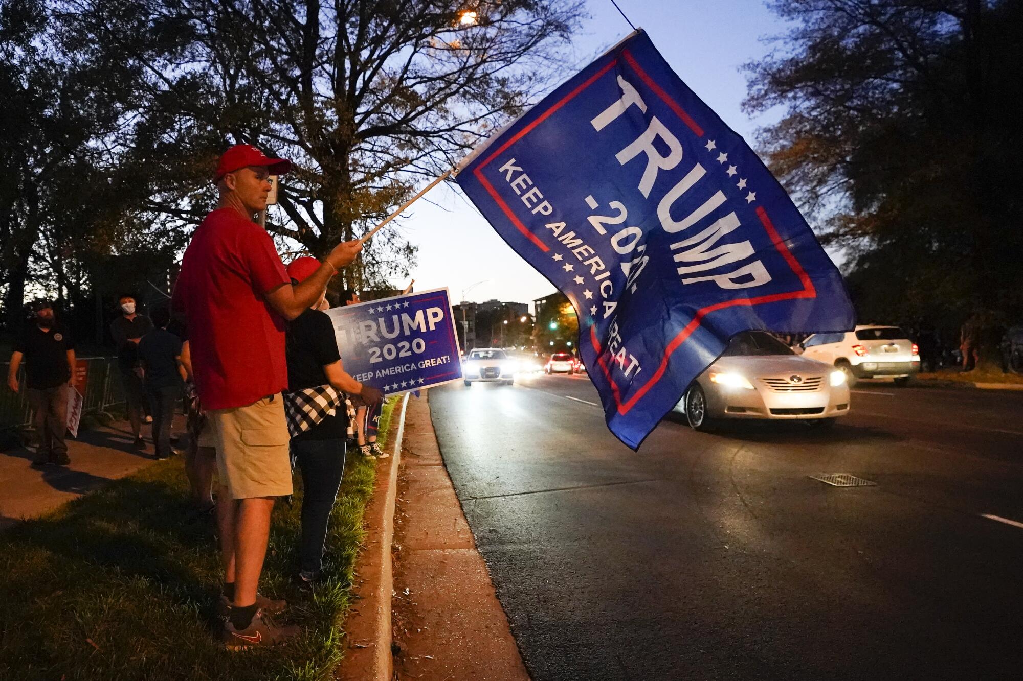 A man holds a Trump 2020 Keep America Great flag on the side of a road as cars drive by