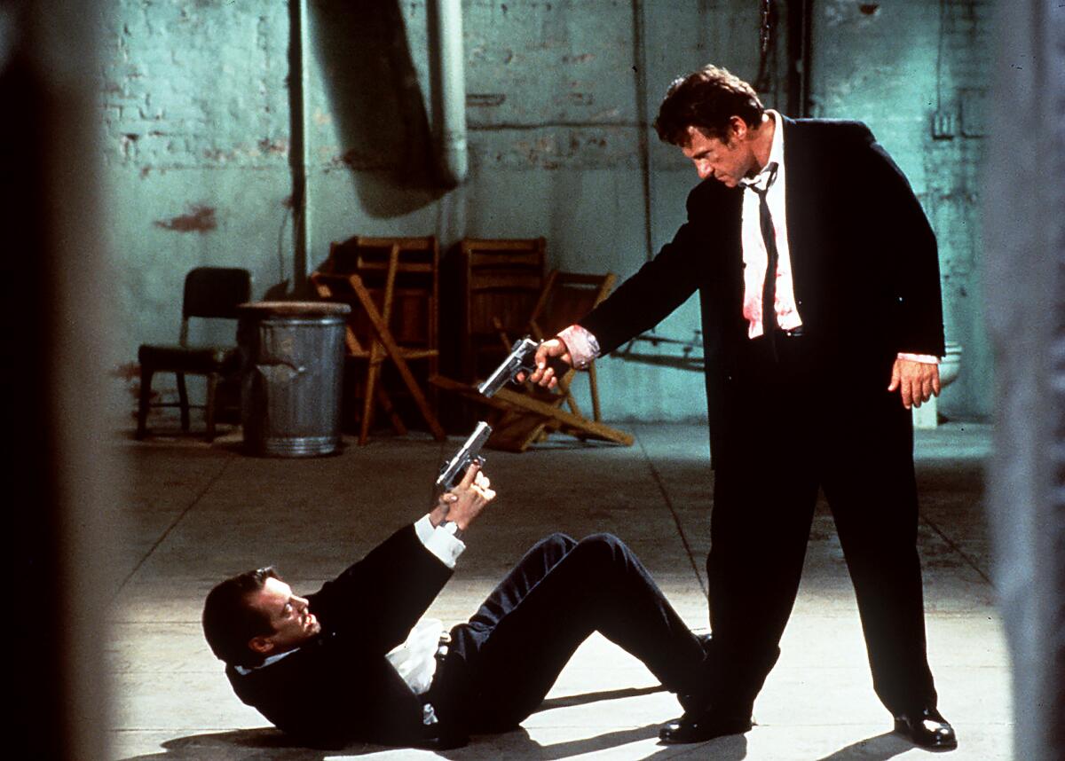 A man on the floor on his back points a gun at the gun-pointing man standing over him