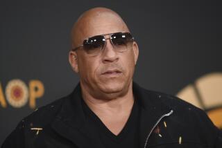 Vin Diesel wearing dark sunglasses and a black formal shirt posing with a straight face against a dark background