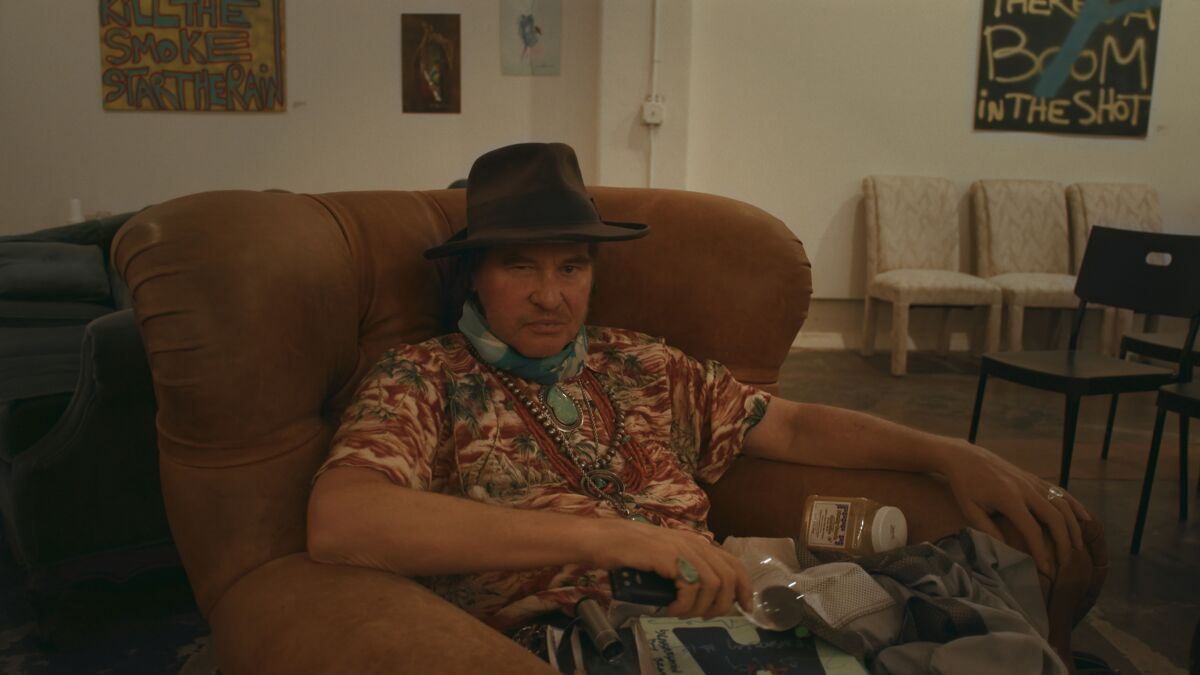 Val Kilmer, wearing a hat and a colorful shirt, sits in a chair.