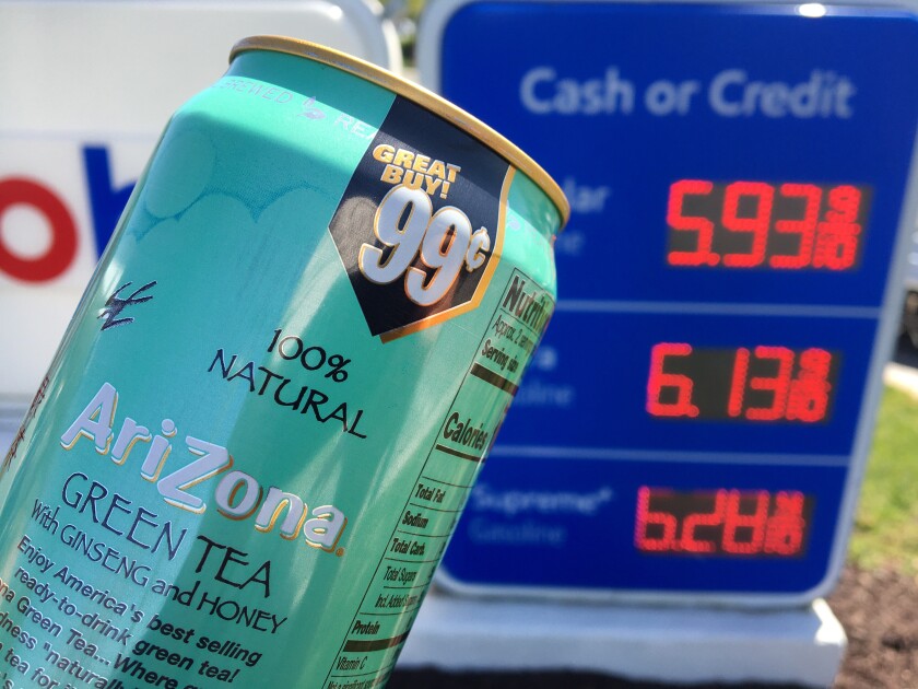 A can of AriZona iced tea in front of a gas station sign displaying high prices.